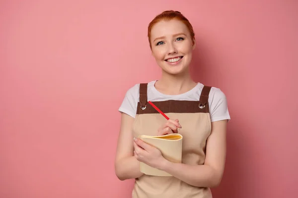 Red-haired girl in an apron with collected hair stands on a pink background with a notebook and smiles broadly. — Stockfoto