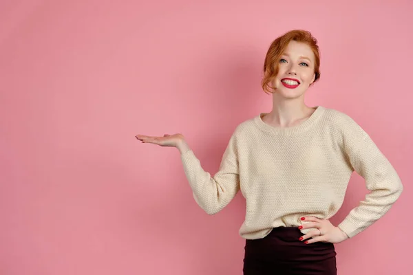 A beautiful redhead girl with red lips in a white sweater stands on a pink background and smiling broadly, points palm to the side — 스톡 사진