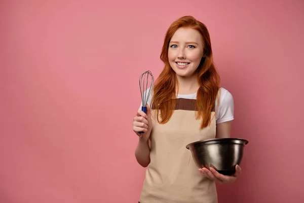 The red-haired girl in an apron stands on a pink background and smiles, holding a bowl and a whisk in her hands. — Stockfoto