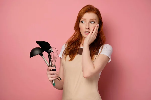 A redhead girl in an apron stands on a pink background with a shovel, a whisk and a ladle in her hand and looks at them in horror — Stockfoto