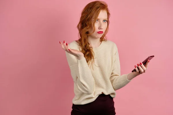 A red-haired girl with red lipstick on a pink background with a phone in hand and is looking upward pursed lips in displeasure — Stockfoto