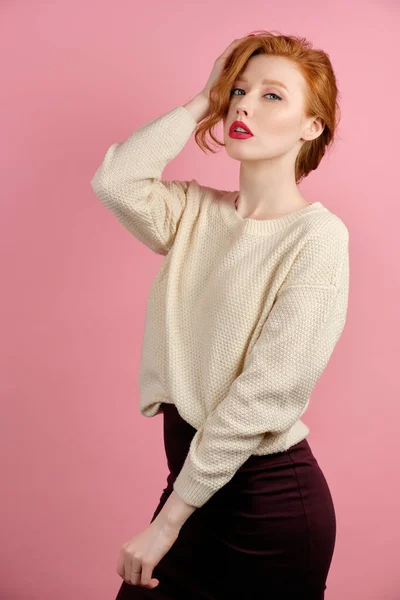 Beautiful redhead girl with red lipstick posing on a pink background and looking at the camera, straightening her hair — 图库照片
