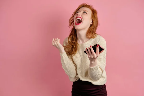 Beautiful red-haired girl with red lipstick screams joyfully up while standing on a pink background with a phone in her hands — Stockfoto
