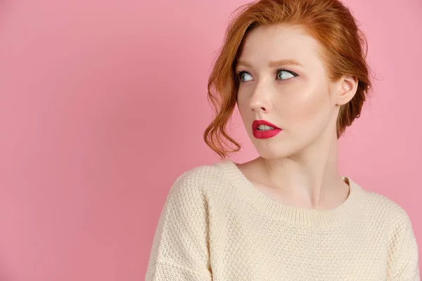 A beautiful red-haired girl with red lips in a white sweater stands on a pink background and, turning her head, looks to the side — 스톡 사진