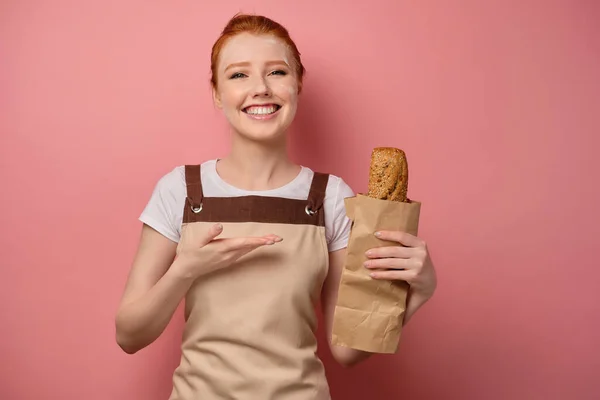 Beautiful red-haired girl with gathered hair in an apron, with flour on her face, smiling, pointing her palm at a bag of bread — Stockfoto