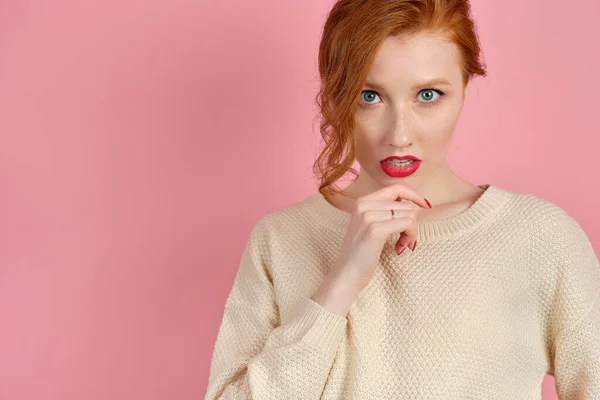 A beautiful redhead girl with red lips in a white sweater looks at the camera, holding chin with fingers — 스톡 사진