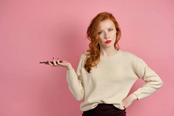 A beautiful red-haired girl with red lipstick stands on a pink background with a phone and looks displeasedly at the camera — 스톡 사진