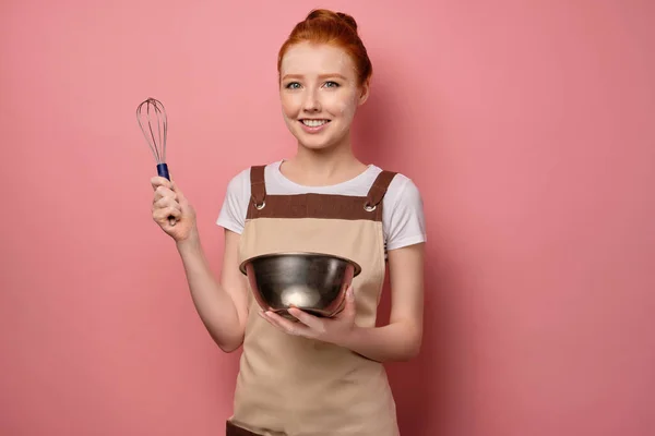Red-haired girl, soiled in flour, in an apron, stands on a pink background with a bowl, holding a whisk up — Stockfoto