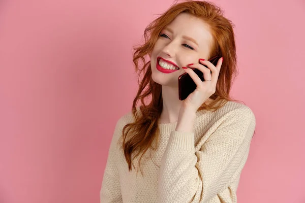 Beautiful redhead girl with red lipstick speaks on the phone while standing on a pink background in half-turn and smiles broadly — 스톡 사진