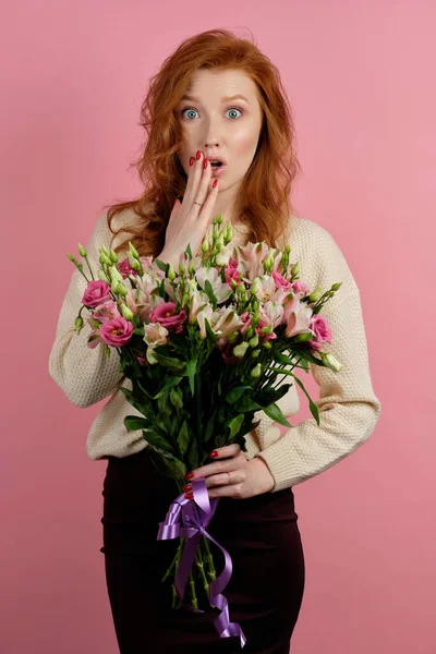 Beautiful redhead girl with a bouquet of flowers on a pink background shockingly covering mouth with hand, looking at the camera — Φωτογραφία Αρχείου