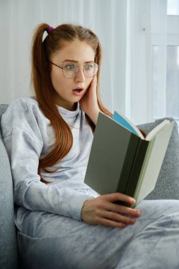 Gözlüklü, uzun kuyruklu, pijamalı kızıl saçlı bir kız kanepede oturur ve kitaba bakar..