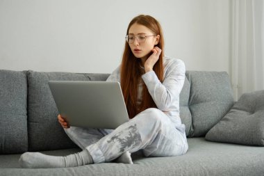 A red-haired girl with glasses, with long hair in pajamas sits cross-legged on the couch and looks at the laptop clipart