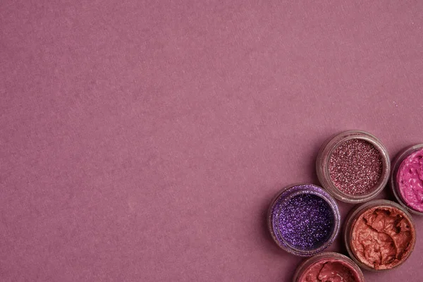 Glitters and lipsticks in lilac colors stand in the corner against a plum textured background