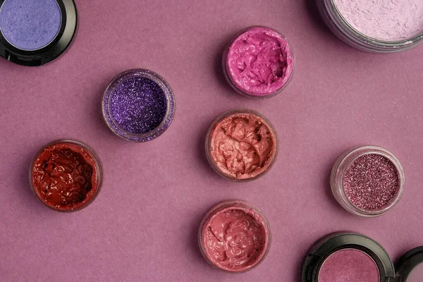 Glitter, lipstick, lip gloss, eye shadow and powder randomly stand on a plum textured background