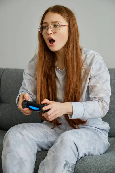 A red-haired girl with glasses, with long tails, sits on a devan with a game console and looks in frustration with her mouth open — Stock Photo, Image