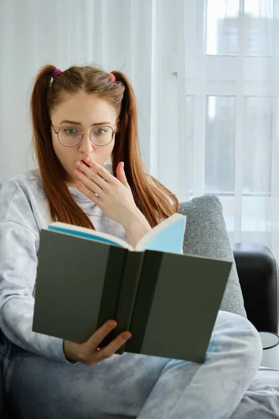 Ruda dziewczyna w okularach, z długimi ogonami w piżamie siedzi na kanapie i patrzy w szoku na książkę, zakrywając usta dłonią — Zdjęcie stockowe