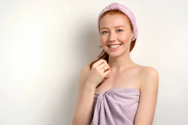 Zrzavá dívka s čistou pletí v ručníku a obvazem na hlavě se široce usmívá do kamery — Stock fotografie