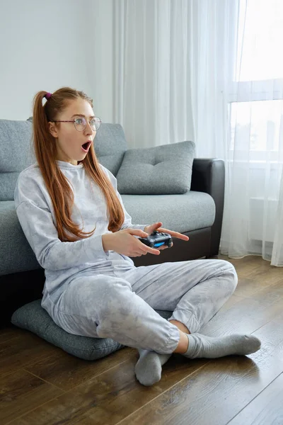 Uma menina de cabelos vermelhos com óculos com caudas longas, senta-se no chão com um console de jogo e abre a boca em choque, olhando para a frente — Fotografia de Stock