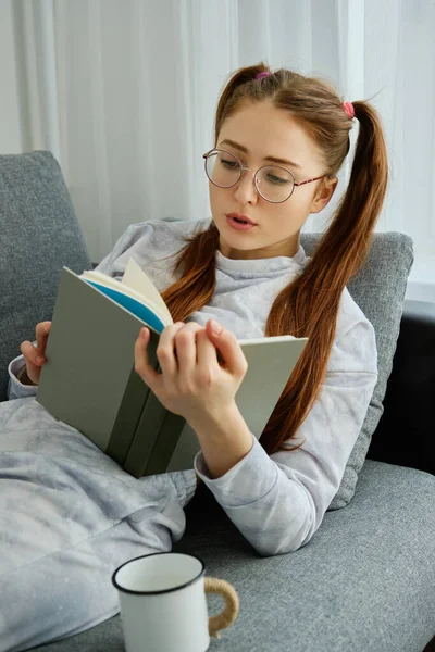 A red-haired girl with glasses, with long tails in pajamas lies on the sofa with a mug and reads a book, focus on her face