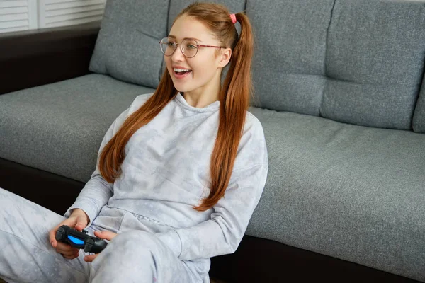 Uma menina de cabelos vermelhos com óculos, com caudas longas senta-se no chão, apoiando-se em um sofá e jovialmente joga em um console de jogo — Fotografia de Stock