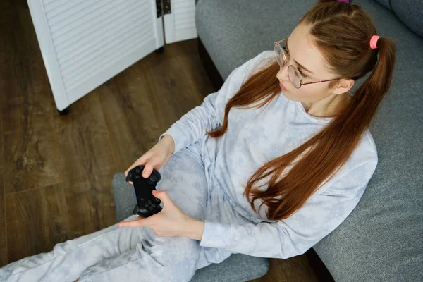 Fille rousse avec des lunettes, avec de longues queues est assis sur le sol dans un demi-tour appuyé sur un canapé avec une console de jeu, tiré par le haut — Photo