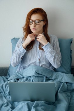Kızıl saçlı, gözlüklü bir kız gömleğinin fermuarını kapatıyor ve dizüstü bilgisayarına bakıyor.