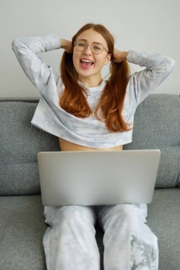 Gözlüklü kızıl saçlı bir kız koltukta dizüstü bilgisayarla oturup saçlarını kuyruklarıyla toplamaktan mutlu bir şekilde gülümsüyor..