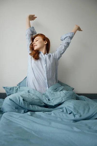 Çizgili pijamalı kızıl saçlı kız yatağa oturur ve ellerini kaldırır.. — Stok fotoğraf