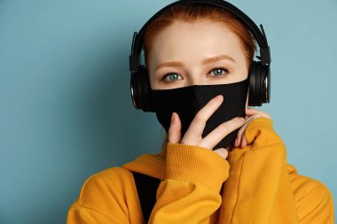 A girl in a yellow hoodie with headphones stands on a blue background and adjusts a black protective mask.  clipart