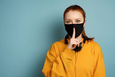 A girl in a yellow hoodie stands on a blue background with her index finger attached to a black protective mask on her face clipart