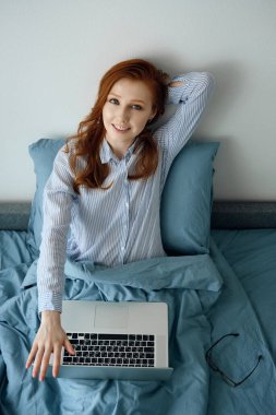 Pijamalı kızıl saçlı bir kız dizüstü bilgisayarla yatakta oturuyor, elini başının arkasına atıyor ve gülümsüyor.