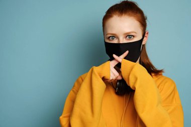 The red-haired girl stands on a blue background in a yellow hoodie crossing her fingers in front of a black protective mask clipart