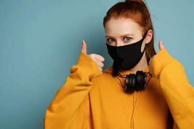 A girl in a yellow hoodie with headphones around neck stands on a blue background in a black protective mask and shows thumbs up clipart