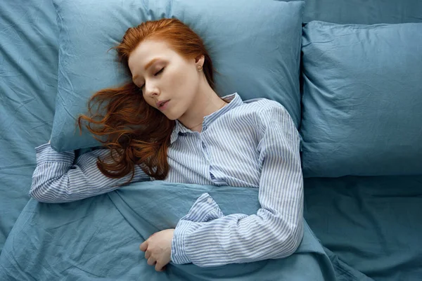 Çizgili pijamalı kızıl saçlı bir kız mavi çarşafların üzerinde, yastıkta uyuyor. Çerçeve üstte — Stok fotoğraf