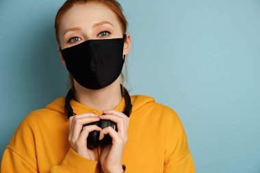 Head Shot. Redhead girl in a yellow hoodie and black protective mask stands on a blue background and looks into the frame clipart