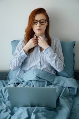 Güzel kızıl saçlı, gözlüklü bir kız yatakta oturmuş, gömleğinin düğmelerini ilikliyor ve dizüstü bilgisayarına bakıyor.