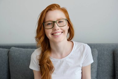 Gözlüklü, kızıl saçlı ve beyaz tişörtlü bir kız çerçevede gülümsüyor.
