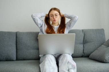 Pijamalı kızıl saçlı bir kız kanepede dizüstü bilgisayarıyla oturur, dudaklarını bir öpücükle gerer ve elleriyle at kuyruğu toplar.