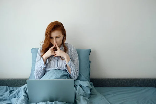 Rudovlasá dívka v pyžamu se zamyšleně podívá do laptopu a přitiskne si prsty ke rtům. — Stock fotografie
