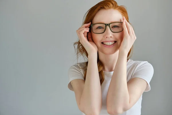 Uma menina ruiva em uma camiseta branca fica em um fundo cinza e sorri enquanto se segura em seus óculos . — Fotografia de Stock