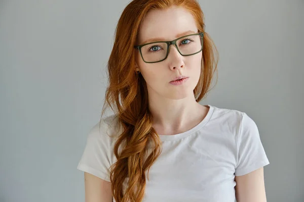 Egy vörös hajú lány fehér pólóban és szemüvegben, elgondolkodva bámul a keretbe, az ajkai a fejét szívják. — Stock Fotó