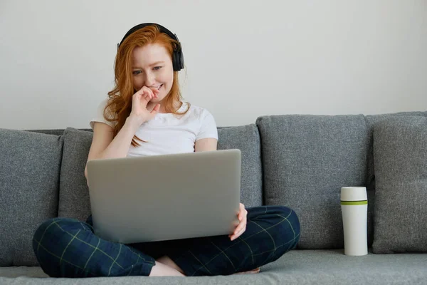 Ragazza rossa in abiti da casa e cuffie si siede sul divano, gambe incrociate e sorridente, guardando il computer portatile — Foto Stock