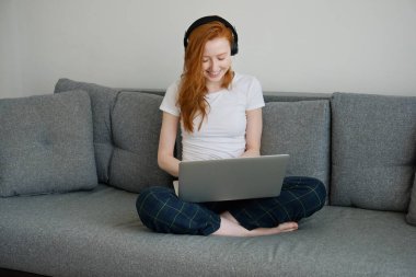 Kanepede oturan kızıl saçlı kız dizüstü bilgisayarla bacak bacak üstüne atmış, gülümsüyor ve aşağıya bakıyor.