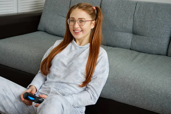 Une fille rousse en lunettes et en pyjama s'assoit sur le sol avec un joystick de jeu et sourit . — Photo