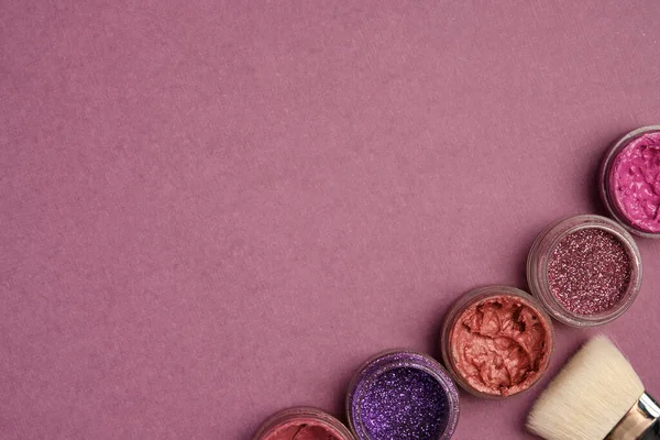 Glitters, lipsticks and a brush in lilac colors stand in the corner against a plum background.