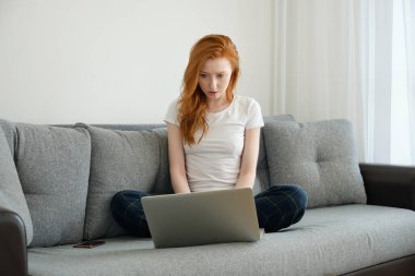 Kızıl saçlı bir kız ev kıyafetleri içinde bağdaş kurmuş oturuyor ve özenle dizüstü bilgisayara bakıyor.