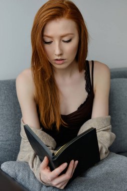 Güzel kızıl saçlı bir kız kanepede oturur ve elindeki açık siyah kitaba bakar.