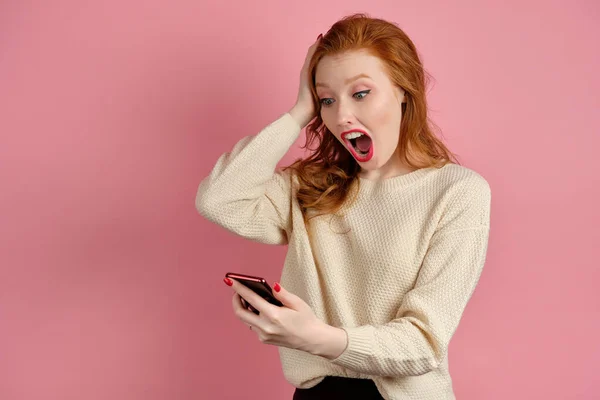 En rödhårig flicka med rött läppstift står i en rosa bakgrund och tittar på telefonen i handen chockad av att öppna munnen och hålla hand på huvudet — Stockfoto