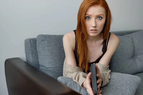 Een roodharig meisje met blauwe ogen zit op een bank met een gesloten zwart boek in haar handen en kijkt in het frame — Stockfoto