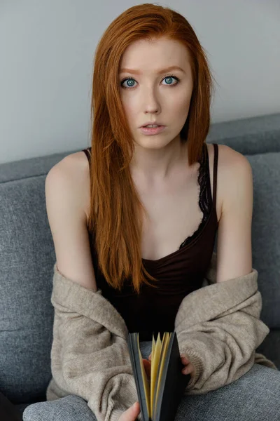 Krásná zrzavá dívka s modrýma očima sedí na pohovce s černou knihou v rukou a dívá se do rámu — Stock fotografie